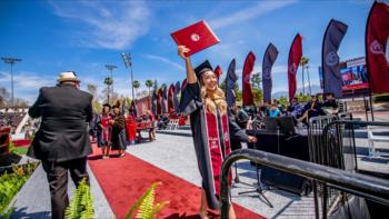 Female student in graduation regalia holding up her diploma as she crosses a stage