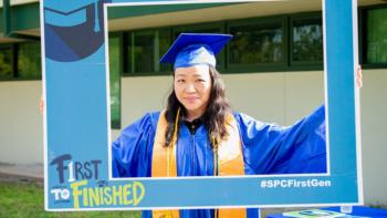 graduate in blue cap and gown holding a 'first to finished' frame
