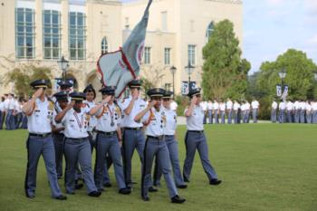 cadets march with a flag on a grassy field in front of a building