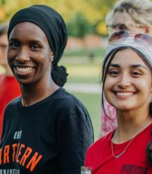 two smiling students at an outdoor event, wearing university shirts