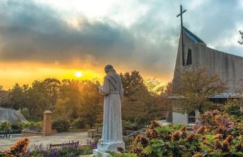statue of a monk with a chapel and cross at sunrise