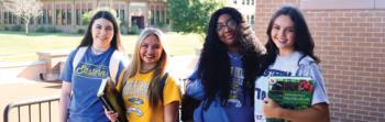 students smiling outside, holding books and wearing college shirts