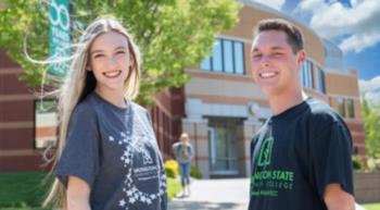 two students smiling in front of a campus building, trees in the background
